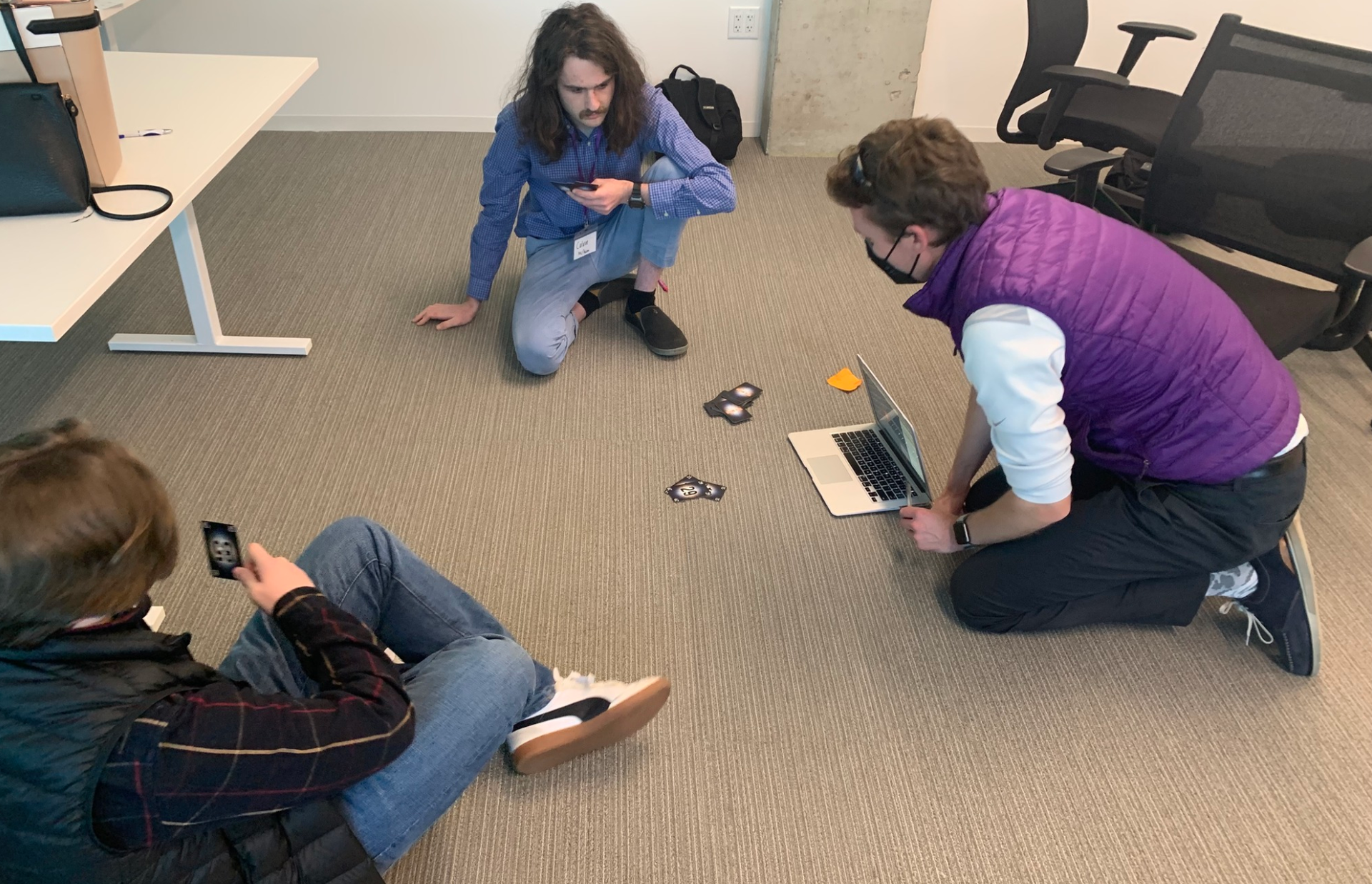 Image shows Game Genius moderator (in purple vest to the right) on the floor talking to two other interns that are seated near him.